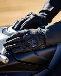 Alpinestars SP X 7 Gloves, black perforated leather motorcycle gloves with TPU knuckles, resting on a fuel tank.