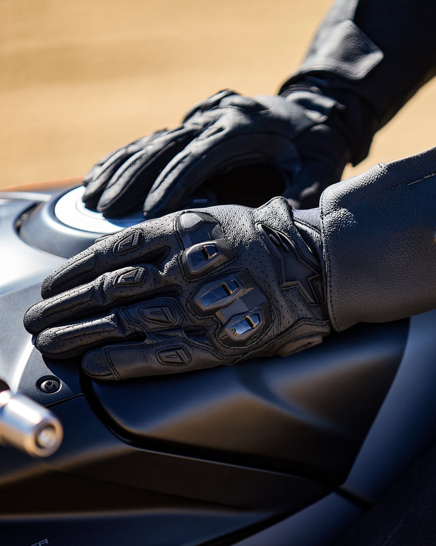 Alpinestars SP X 7 Gloves, black perforated leather motorcycle gloves with TPU knuckles, resting on a fuel tank.