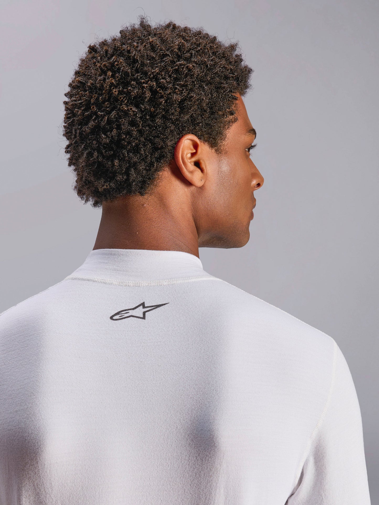 Alpinestars ZX Evo V3 Top, Long Sleeve Auto Racing Base Layer, White, close-up of back neck area featuring black Alpinestars logo, technical flame-resistant fabric, worn by model with dark curly hair